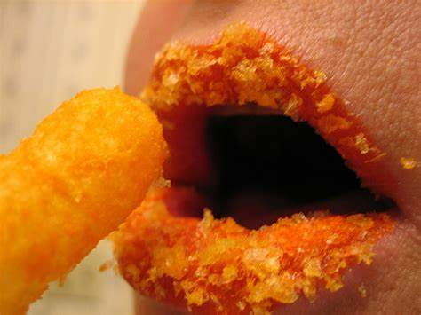 A close-up of open lips with a Cheeto puff moments before being eaten. Cheeto crumbs densely line the lips, waiting patiently.