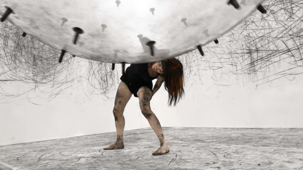 Dancer interacts with a sphere covered in charcoal sticks, leaving marks on the gallery’s white walls and on the dancer's body, at the interactive installation ADA.