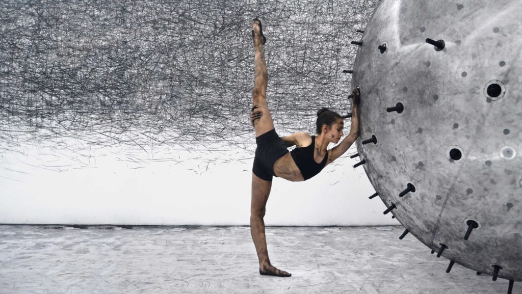 Dancer interacts with a sphere covered in charcoal sticks, leaving marks on the gallery’s white walls and on the dancer's body, at the interactive installation ADA.