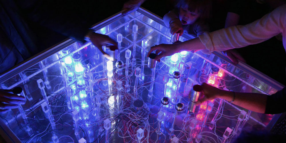 Several visitors’ hands moving metal balls by a magnetic pawn on the mirrored and illuminated, in the interactive installation The Brain Game.