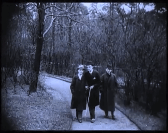 Looped black and white gif of the same two white mascs in suits, coats, and hats walking arm-in-arm in a park, one holding a cane; another white masc sidles up to them and says "handsome lad!" while leaning close; the older of the couple responds angrily while the other clutches his chest in offense and confusion; they walk away. 