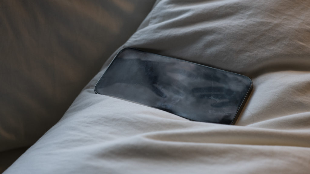 An iphone with a faint image of two hands touching sitting on fabric that appears to be bed sheets.