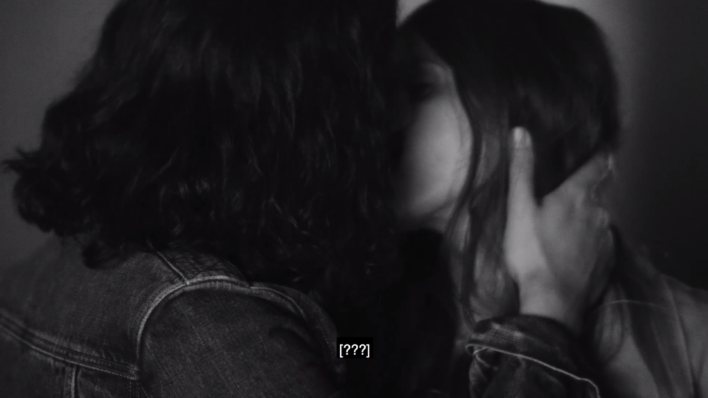 A black and white film still of a Caucasian man and woman kissing. The man has his right hand on her neck.  The subtitles read “[???].” They both have long black hair. He is wearing a dark denim jacket.