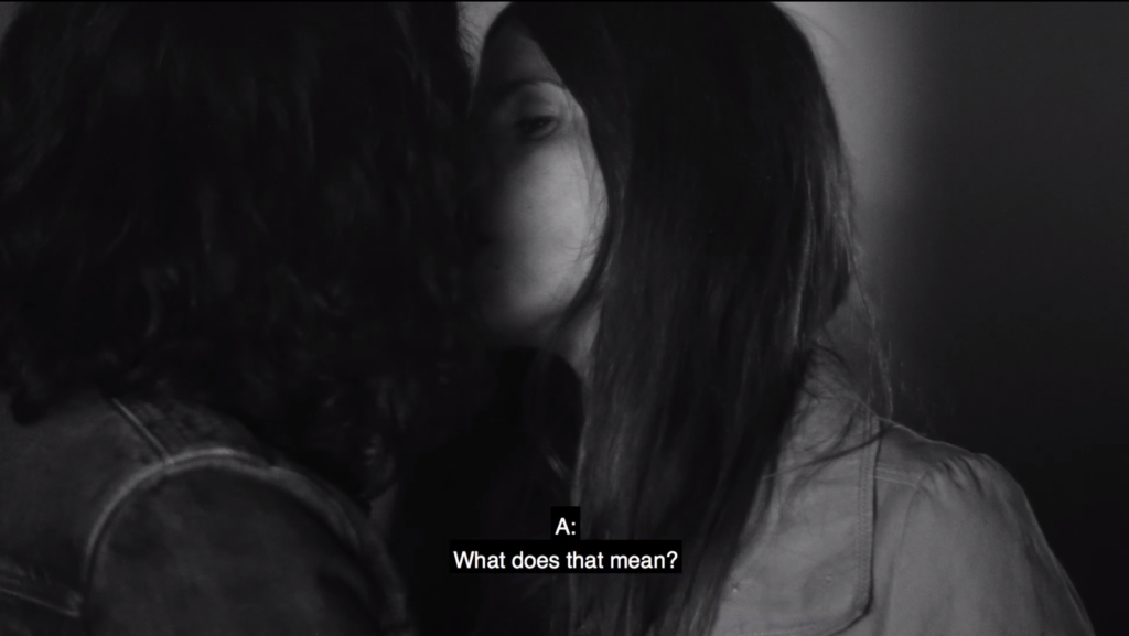 A black and white film still of a man and woman kissing. The subtitles read: “What does that mean?” She is wearing a light colored trench coat.