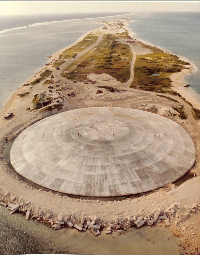 The Runit Dome (or Cactus Dome), in the foreground, a large cement dome sitting on a small island just barely bigger than the dome itself. The ocean can be seen all around with some scrub brush on the island as well.