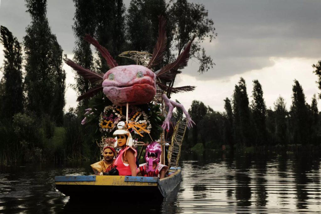 A boat flows through a river before a background of trees. In the boat, a figure dressed in pink and wearing a gold helmet looks towards the foreground. The figure is flanked by two seated figures dressed in yellow and pink. A large sculpture of an axolotl balanced on a structure covered in flowers and vines is above the figures.