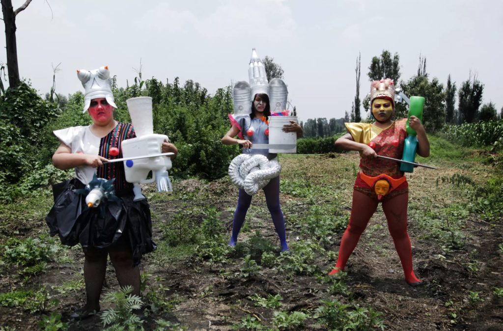 Three figures stand in a crop field, their legs spread hip-width apart. They each wear items resembling armor, including breastplates, helmets, and drum-like objects, made out of plastic jugs, and other assembled items.