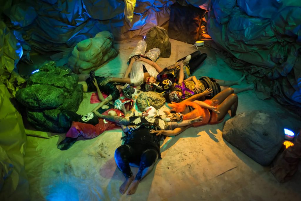 Seven figures dressed in bright-colored clothing and metallic helmet-like headpieces lie in a circle on the ground of a cave. Blue and green metallic fabric is bunched around the edges of the scene alongside a few large rocks. Each figure has their arms outstretched to touch the torso of the figure next to them.