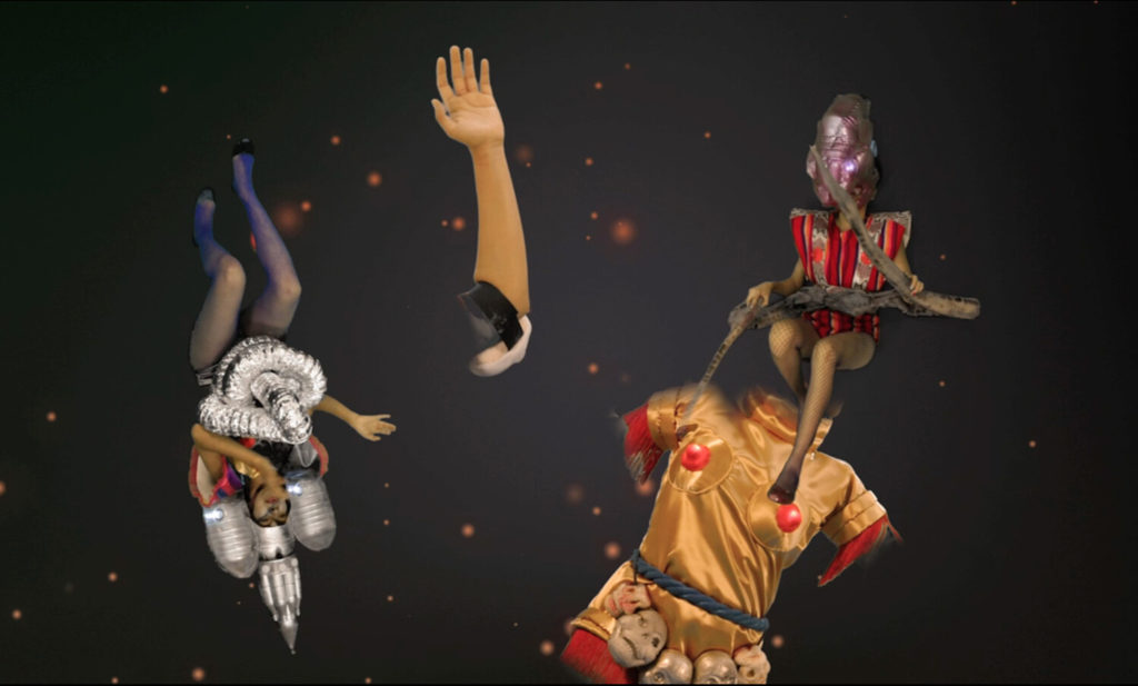 Against a dark background with glowing orange dots, three figures float around a disembodied arm. The figure on the left is upside down, wearing blue tights and a metallic set of chest armor, and a pointed helmet. To the right, another figure wearing red holds a long twisted brown object that appears like a branch or a snake. The figure’s face is covered by a large purple metallic helmet. Below, a large torso of a figure is seen wearing a gold tunic with a belt made of skulls. Their chest is protected by two circular pads, each one with a red center covering the figure’s nipple.