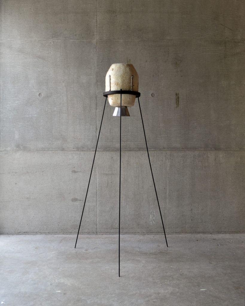 A studio photo of the mycelium sculpture, an off-white cylinder with metal components, held in a steel tripod about 5 feet tall.